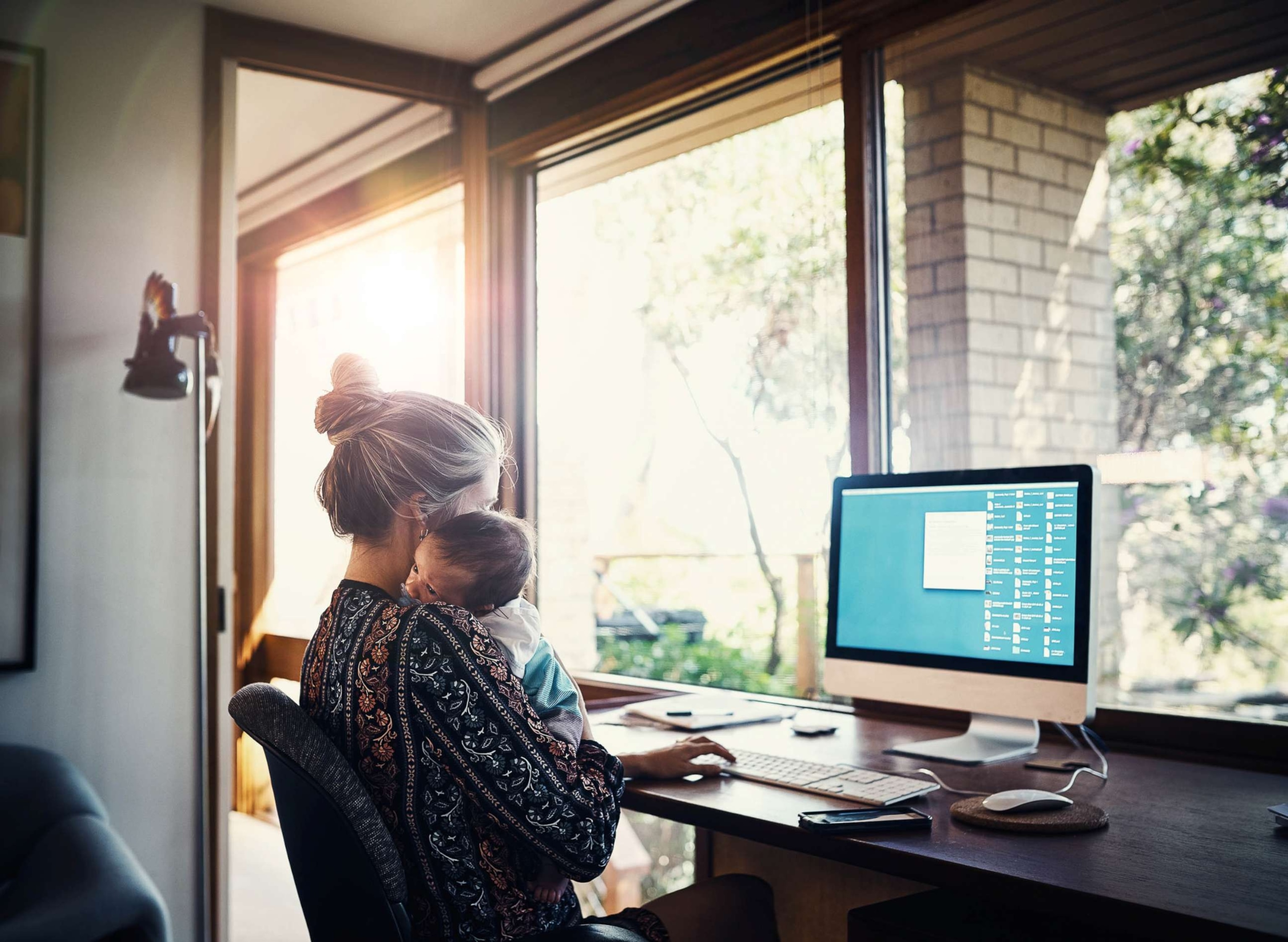 A mother works on a laptop at home while holding her newborn baby.