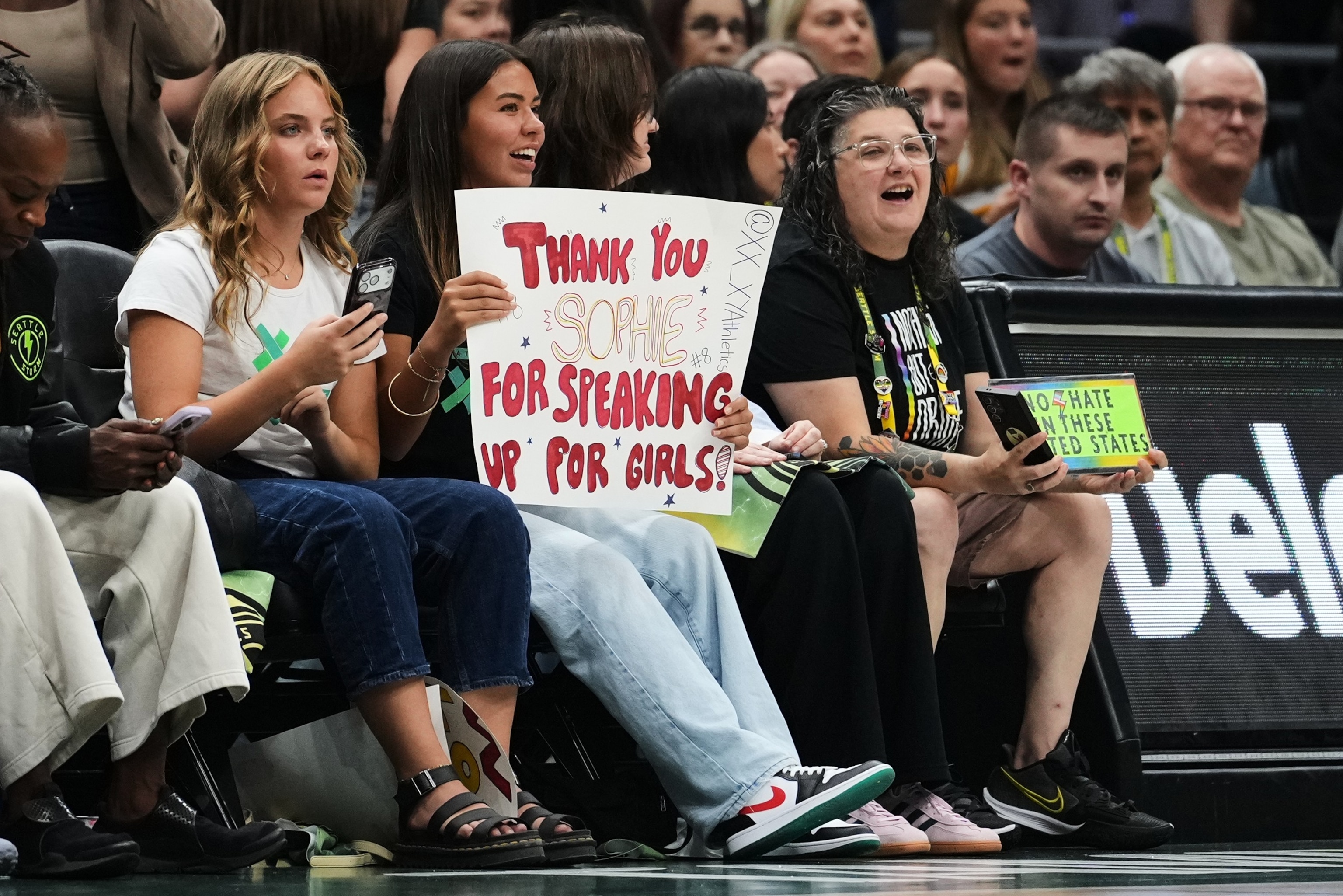 PHOTO: A fan holds a sign for Indiana Fever guard Sophie Cunningham in reference to her comments on transgender athletes as a fan holds a competing sign during the first half of a WNBA basketball game, July 28, 2026, in Seattle. 