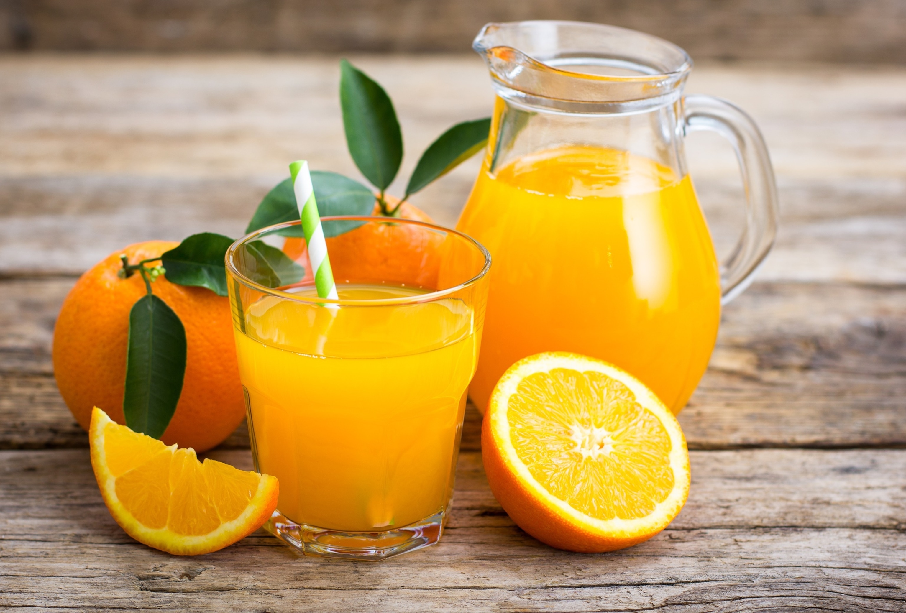 This image showcases a vibrant and refreshing scene centered around freshly squeezed orange juice. The composition is set on a rustic wooden table with a clear glass pitcher and a glass of orange juice surrounded by whole and sliced oranges.
