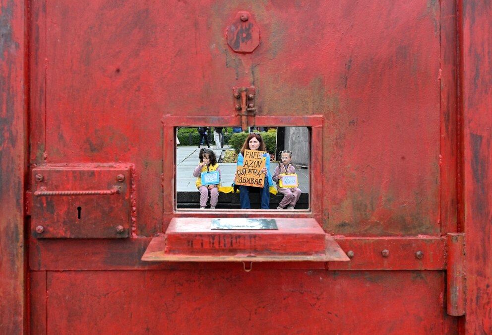 PHOTO: A protester and two children hold placards as they are seen through a window of "door to a prison cell" installation during a rally marking third anniversary of the capture of Azov Brigade fighters in Mariupol, in Kyiv on May 18, 2025.