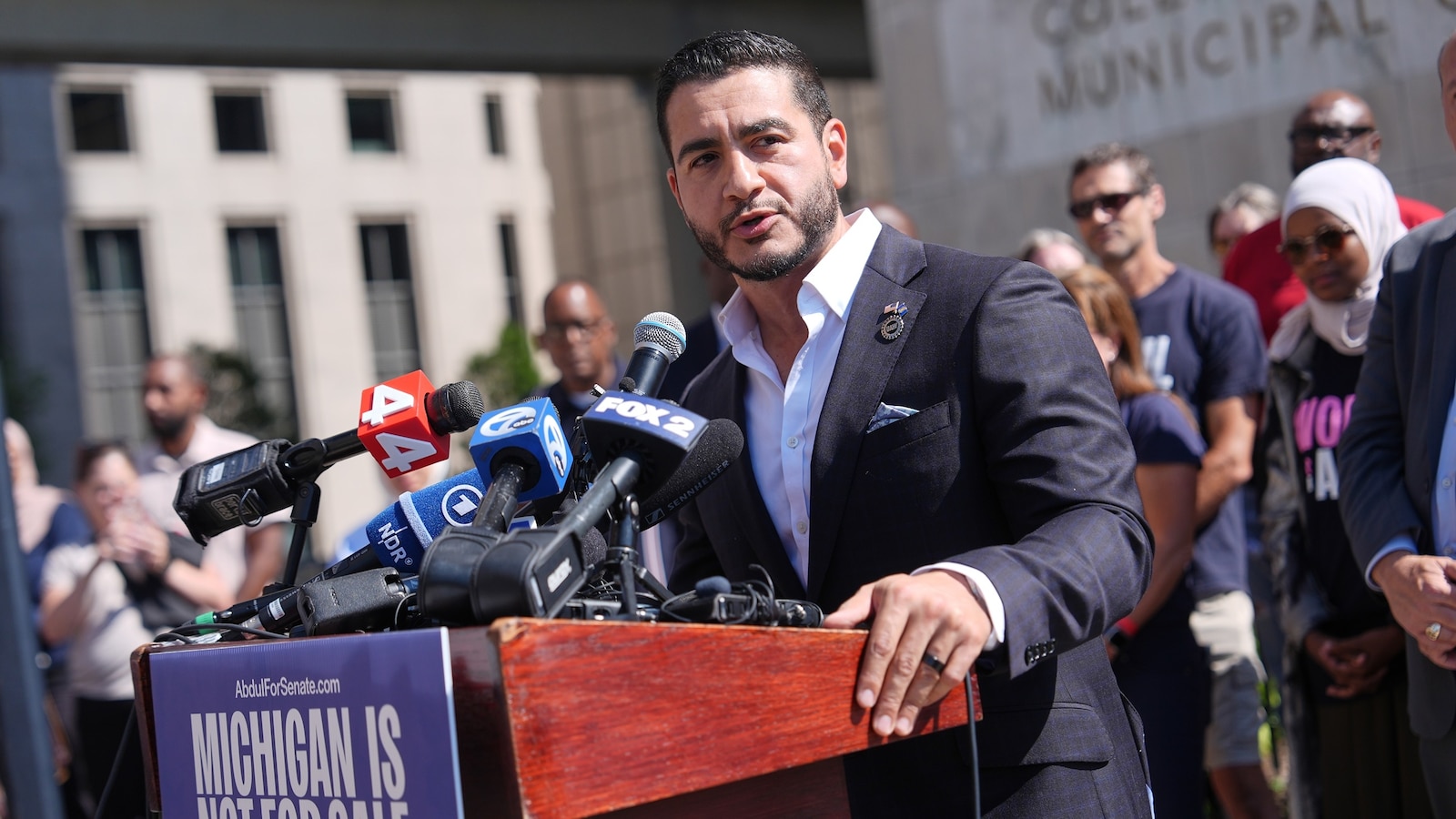 A man in a suit speaking at a podium surrounded by microphones.