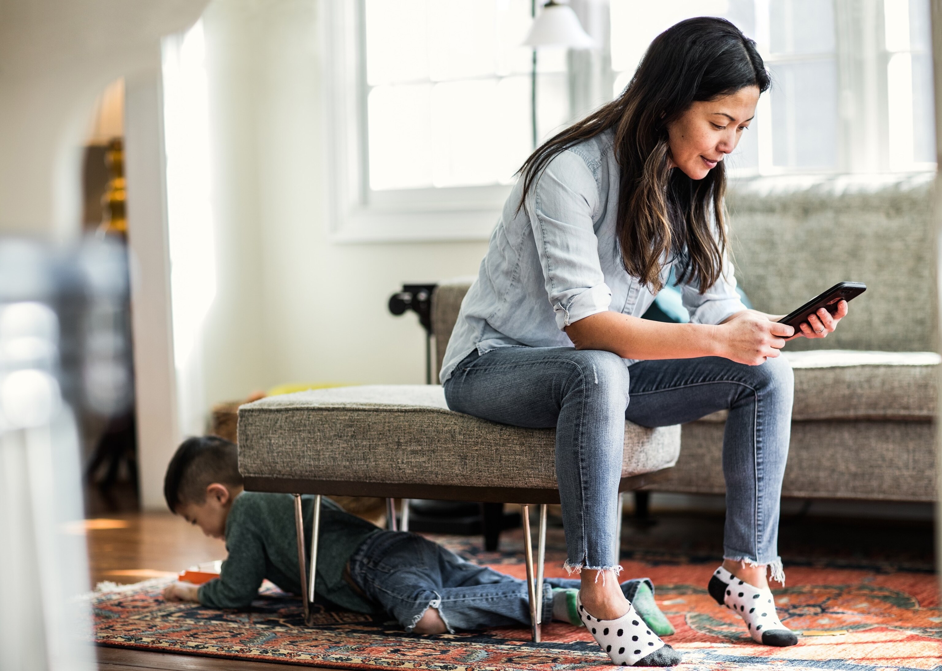 A woman uses her smartphone in the foreground while her child plays in the background.
