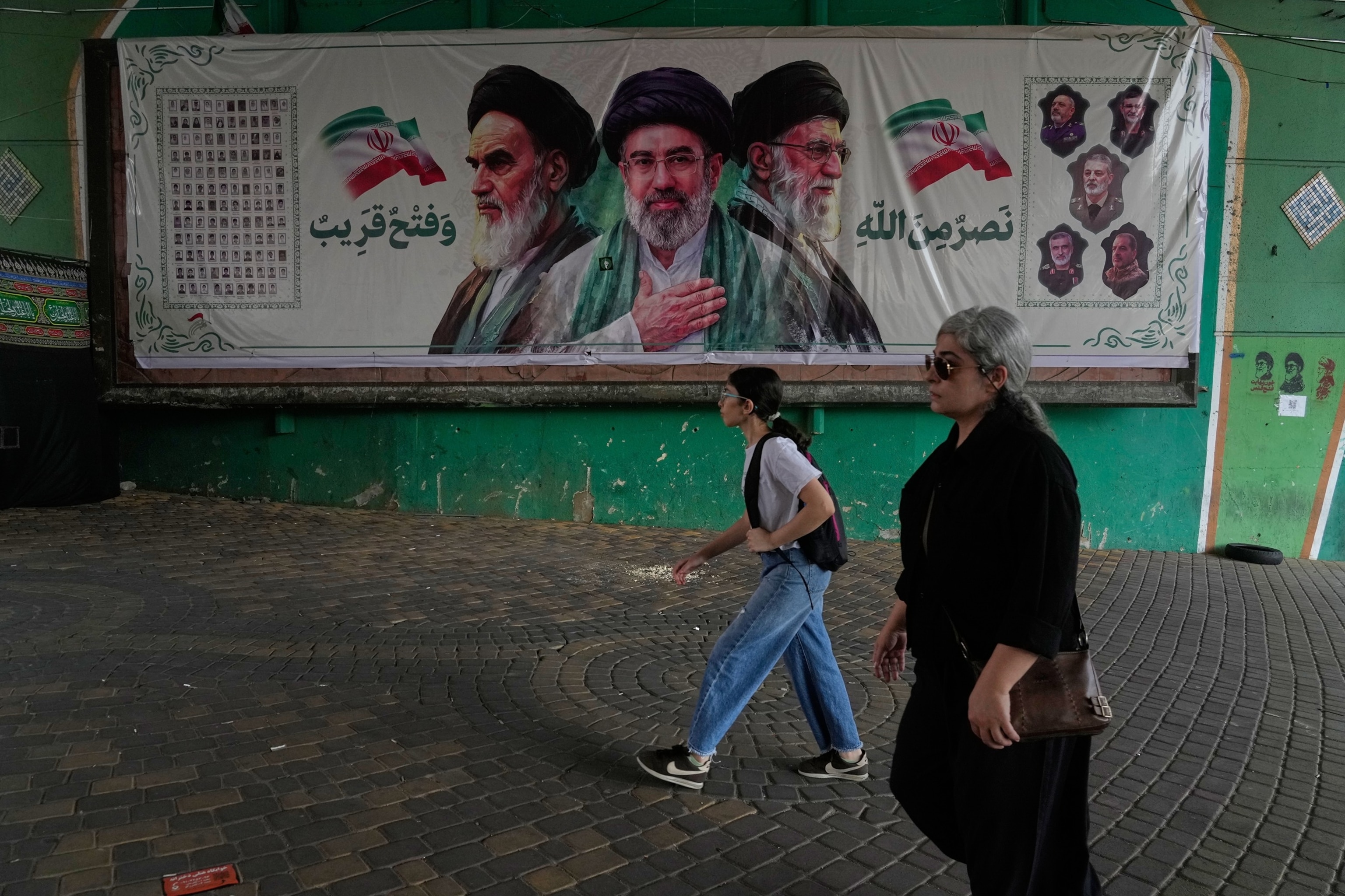 PHOTO: Women walk past a banner showing portraits of Ayatollah Ruhollah Khomeini (L), Supreme Leader Ayatollah Mojtaba Khamenei (C), and the late Supreme Leader Ayatollah Ali Khamenei, under a bridge in northern Tehran, Iran, on Aug. 2, 2026.