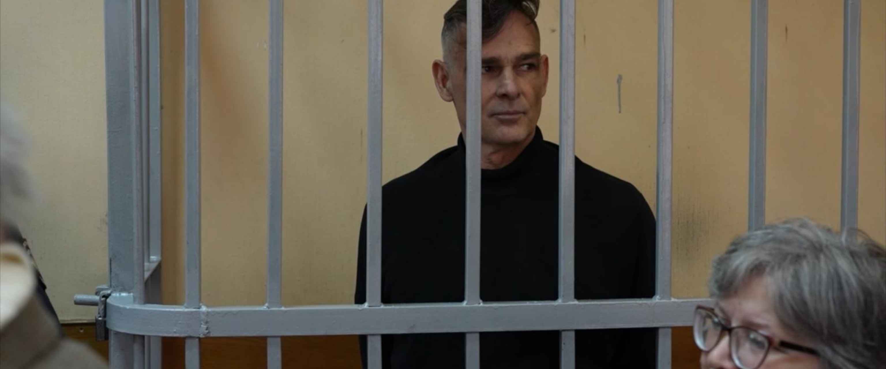 A man with short dark hair, wearing a black turtleneck, is standing behind vertical metal bars that appear to be part of a holding cell or cage. He is looking slightly to his right with a neutral expression on his face. In the foreground to the right, a woman with short gray hair and glasses is partially visible, her face turned slightly downward. The background is a plain beige wall.