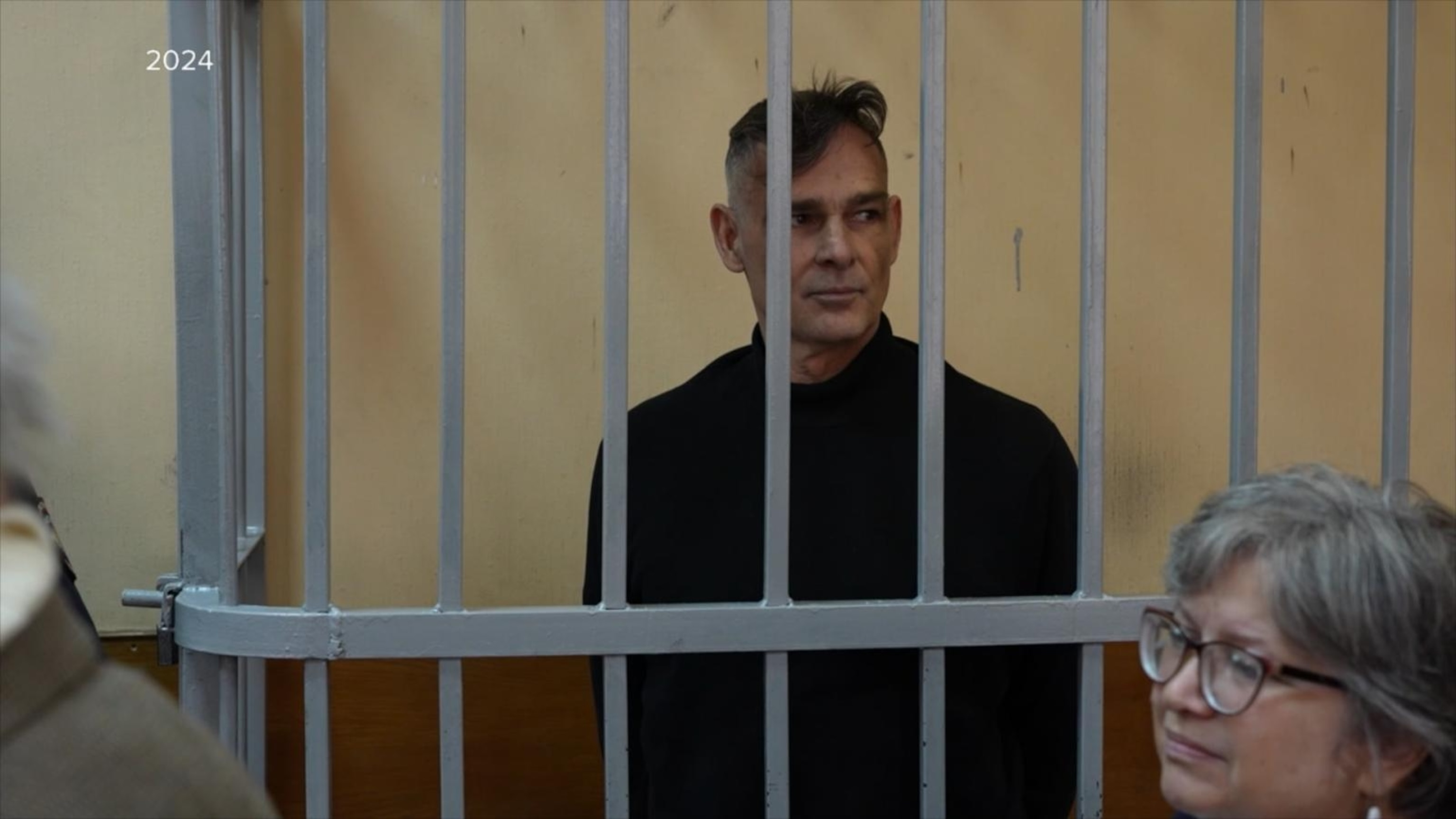 The image shows a man standing behind metal bars in what appears to be a holding cell or courtroom cage, suggesting a detention or legal setting. He has short dark hair with a slight wave and is wearing a black turtleneck sweater. In the foreground, slightly out of focus, is a woman with gray hair and glasses, facing to the right and looking concerned or thoughtful. The walls behind the man are a plain beige color, and the year 2024 is visible in white text on the left side of the image.
