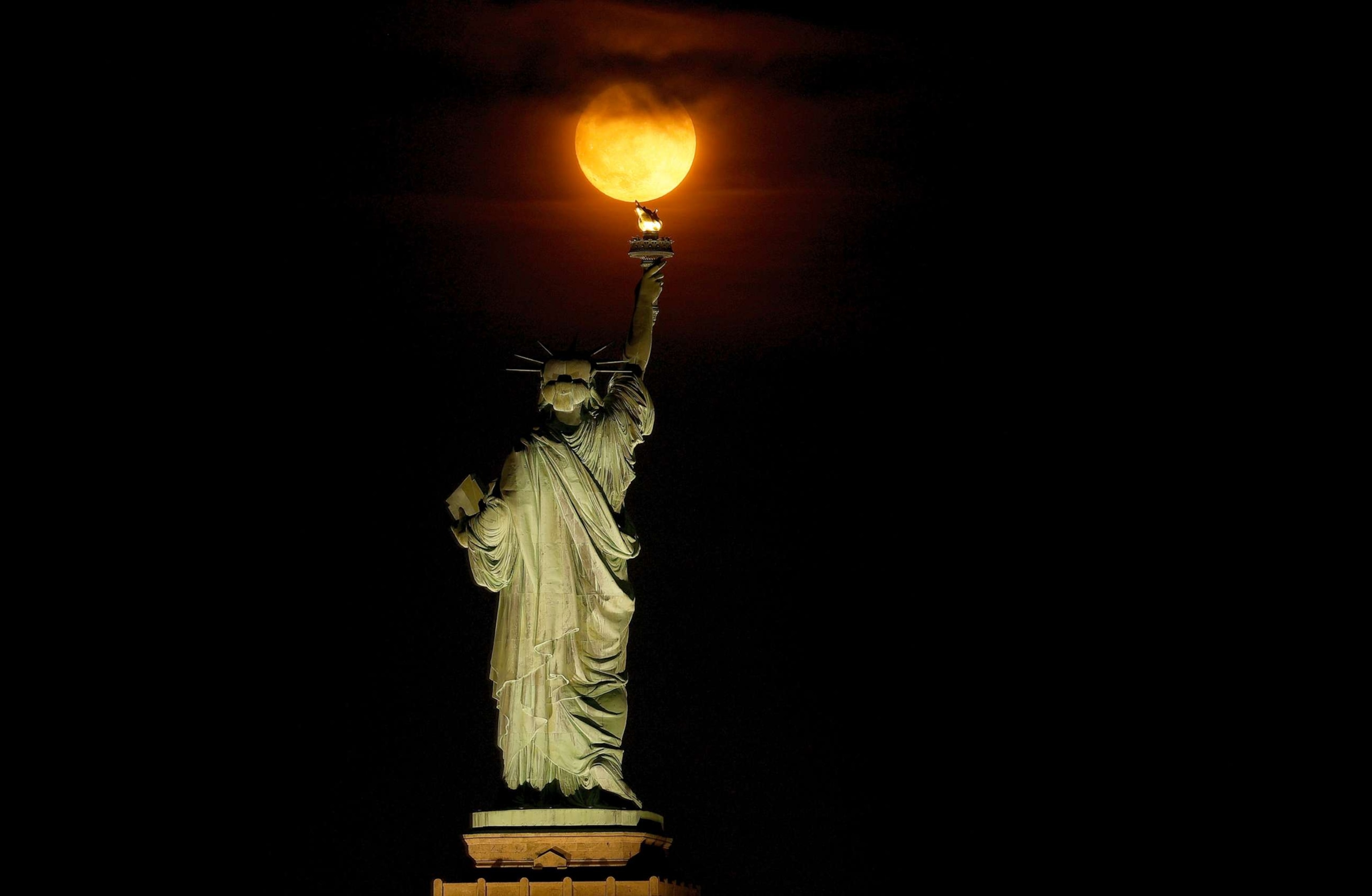 PHOTO: The Buck Moon rises through a haze behind the Statue of Liberty in New York, July 2, 2023, as seen from Jersey City, N.J.