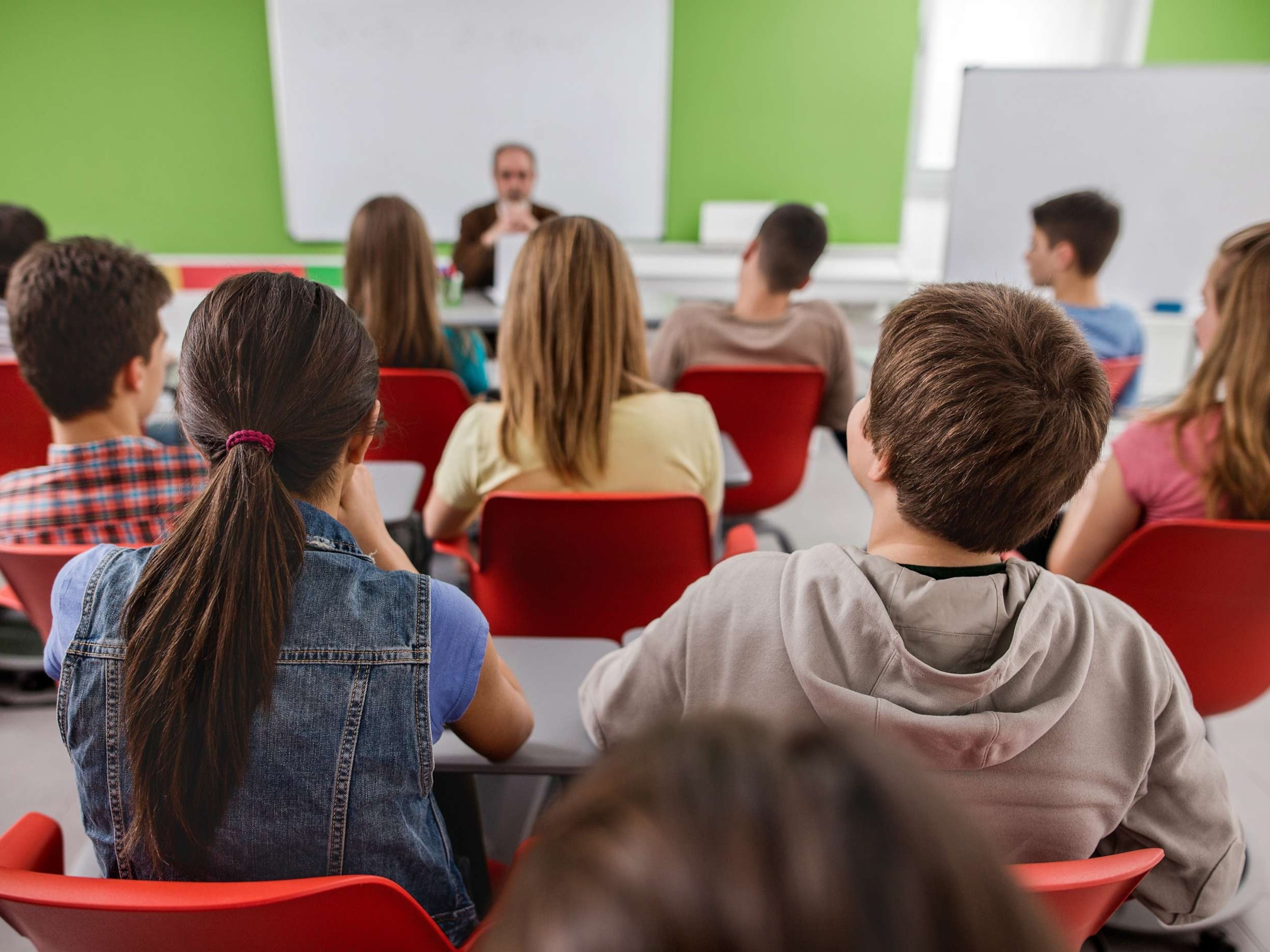 PHOTO: Back view of large group of students sitting in the classroom and listening to their teacher.