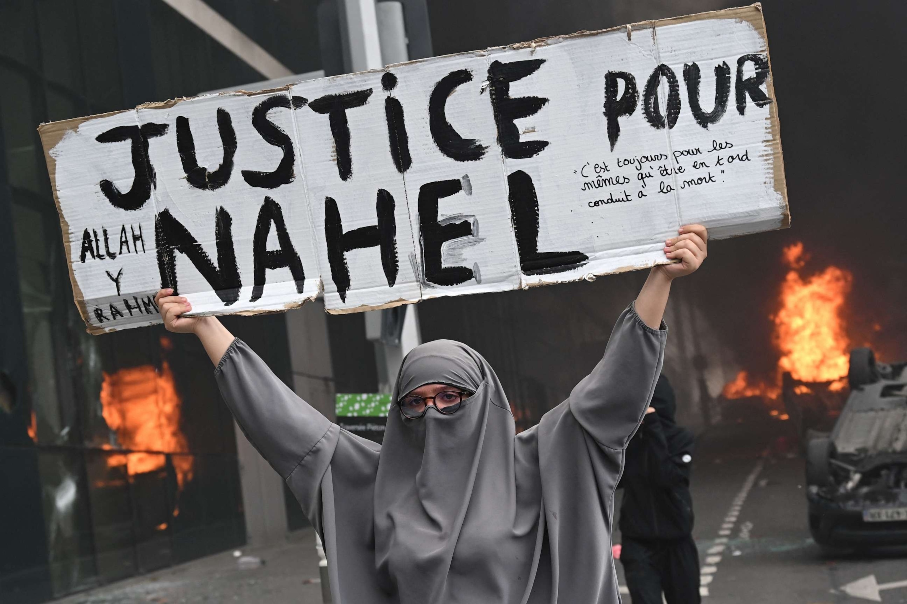 PHOTO: A protester holds a banner reading "Justice for Nahel" as cars burn in the street in the Paris suburb of Nanterre, France, on June 29, 2023, at the end of a commemoration march for a 17-year-old driver who was shot dead by a police officer.